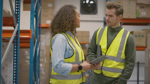 Male and female workers with digital tablet in distribution warehouse meeting and talking surrounded by shelves - shot in slow motion