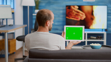 Back view of man holding digital tablet with green screen watching social media video content sitting on sofa. Person looking at touchscreen device with chroma key looking at influencer vlog.