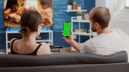 Couple holding vertical smartphone with green screen talking about social media content in home living room. Man and woman using mobile phone with chroma key in online conference or group video call.