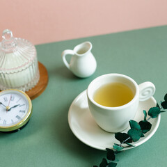 Cup of tea, clock, candle, eucalyptus leaf on green table. pink wall background