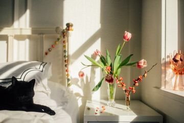 black cat on white bed with pink flowers in vases bedside