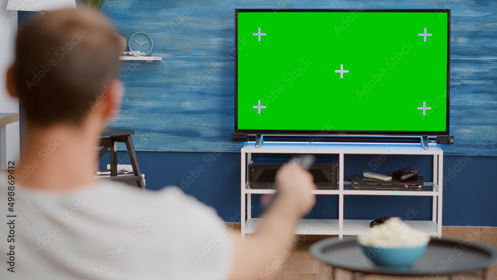 Poster over shoulder view of man switching channels while looking at green screen on tv and sitting on sofa