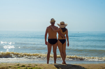 Man and woman in swimsuits and straw hats enjoy the view of the sea on the beach. A beautiful couple standing on a tropical beach and enjoys the view to the turquoise ocean during their summer holiday