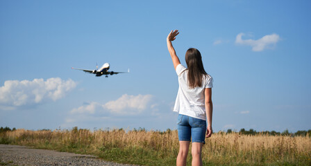 Back view of woman waving hand to flying commercial airplane in the sky. Lifestyle and travel concept.