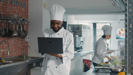 African american cooker looking at laptop screen to cook gourmet dish, following online food recipe on computer. Authentic male chef preparing meal ingredients for gastronomic cuisine menu.