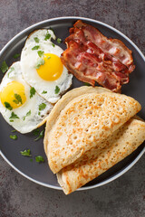 Traditional English Staffordshire oat pancakes, bacon and fried eggs close-up in a plate on the table. Vertical top view from above