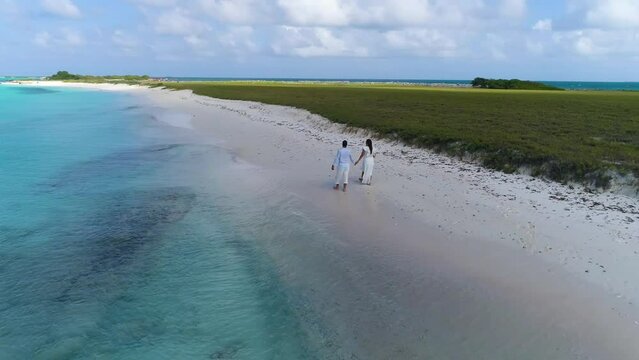 Couple walking on white clothes and barefoot on white sand caribbean beach in Los Roques