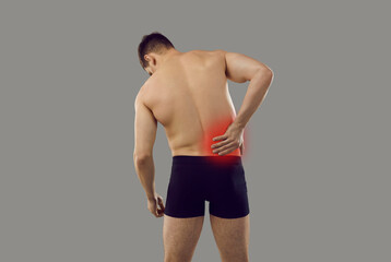 Fototapeta na wymiar Backside of young man suffering from pain in right side. Patient in underwear standing isolated on grey background and holding hand on his inflamed lower back, rear view from behind. Backache concept
