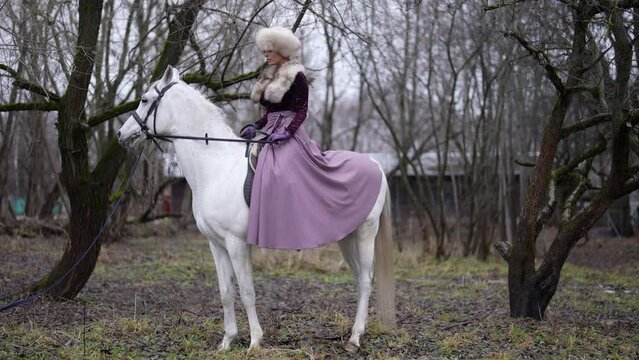 history atmosphere, woman in fur clothing is riding white horse in forest in autumn or winter day