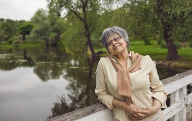 Fototapeta na wymiar Smiling mature grey-haired woman relax on lake enjoy leisure weekend outdoor. Happy elderly grandmother stand on wooden bridge near river dreaming thinking of good maturity or retirement.