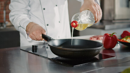 Authentic cook adding organic olive oil in frying pan, preparing hot liquid to fry vegetables and...