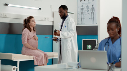 Fototapeta premium Physician giving bottle of pills to expectant patient at checkup, preparing prescription treatment for woman with baby bump in cabinet. Male doctor consulting person at appointment.