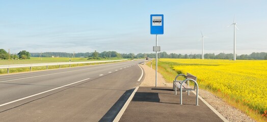 Bus stop and country highway (new asphalt road) through the green forest meadow and blooming yellow rapeseed field at sunrise. Fog, soft sunlight. Transportation, remote places, vacations concepts - 498874485