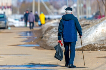 An elderly man walks along the city street with a walking stick, carries a package of groceries...