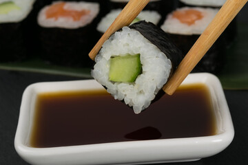 chinese chopsticks hold a roll with fresh cucumber over a cup of soy sauce on a dark background