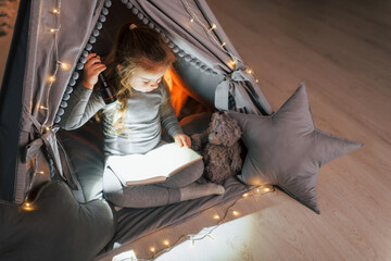 Obraz na płótnie Canvas Reading book at evening. Cute little girl playing in the tent that is in the domestic room