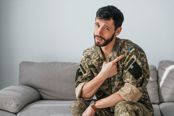 Fototapeta premium Soldier in uniform is indoors in the domestic room. Sitting on the sofa