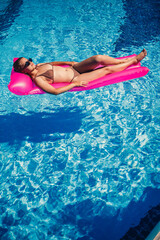 Sexy woman in a swimsuit lies on a pink inflatable mattress in the pool. Relax by the pool on a hot summer sunny day. Vacation concept
