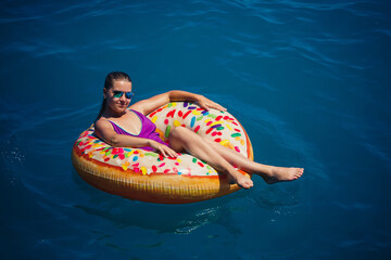 A young beautiful girl in a bright swimsuit lies on a large inflatable ring and floats on the blue sea on a bright sunny summer day