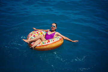 A young beautiful girl in a bright swimsuit lies on a large inflatable ring and floats on the blue...