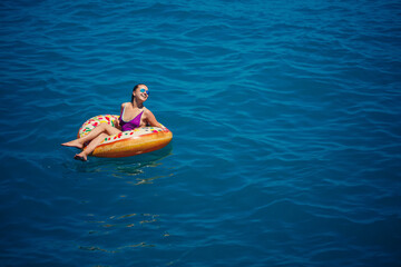 A young beautiful girl in a bright swimsuit lies on a large inflatable ring and floats on the blue sea on a bright sunny summer day
