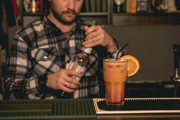 Glass with Tequila Sunrise cocktail on bar counter on background of bartender closing bottle