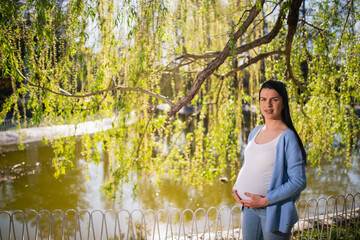 The pregnant woman poses for the camera in a park near the lake. A nice-looking smiling caucasian holds her hand under her belly. There are branches with leaves in the background. 