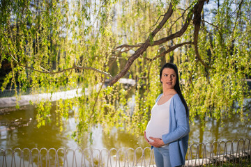 Well-disposed black-haired pregnant caucasian woman stands near lake in park. Beautiful, cheerful female holds her tummy with both her hands and looks at left side. Branches with leaves are behind her