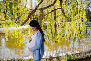 Young caucasian woman in advanced pregnancy cuddles her tummy with both her hands and looks at it.Black-haired pregnant female stands beside lake under the tree with long branches full of green leaves