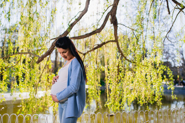 An advanced pregnant woman stands in a park near the lake under the tree, looks at her big tummy, and holds her belly with both hands. Happy female enjoys spring day.