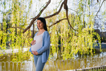 A black-haired well-looking caucasian pregnant woman stands beside a lake in a green park and looks ahead while she cuddles her big belly. A smiling child-bearing female enjoys spring day. 