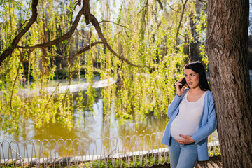 Good-looking, black-haired caucasian child-bearing pregnant woman has a phone call in green park. She leans against tree near lake and looks thoughtfully somewhere while she speaks. She touches tummy.