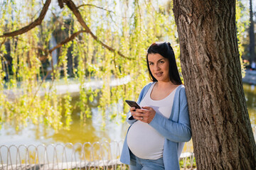 A beautiful pregnant female in 9th month of pregnancy looks at the camera, smiles, and holds her smartphone. She leans against the tree full of green leaves.Lake is in the park. She is beside the lake