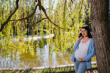 Attractive pregnant woman makes selfies with her smartphone while she is leaning against tree in park. Her left hand is on the belly while she keeps the mobile phone up. Caucasian is next to the lake.