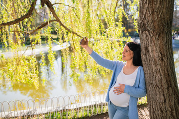 Attractive pregnant woman makes selfies with her smartphone while she is leaning against tree in park. Her left hand is on the belly while she keeps the mobile phone up. Caucasian is next to the lake.