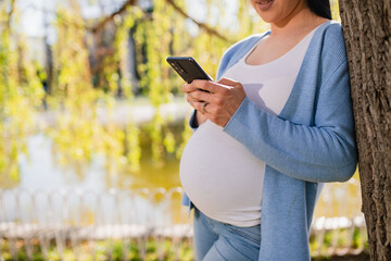 Unrecognizable pregnant caucasian woman types on her smartphone while she leans against the tree in forest. Casual clothed female has wedding ring on her left hand. Lake and branches are behind her. 