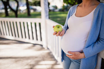 An unrecognizable handsome pregnant female leans against a white fence in the pavilion. A green apple is in her right hand and she holds her belly with her left hand. A woman is casually clothed. 