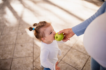 A cute caucasian little girl with ponytails stands and eats a green apple while pregnant...