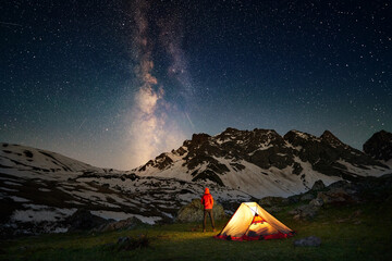 Hiking tourist standing near illuminated tent under milky way stars - 498890488
