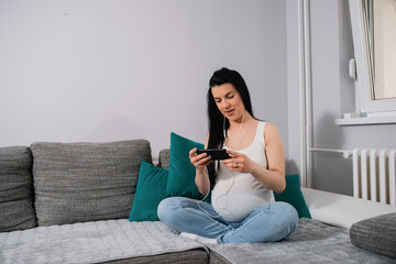 An expectant mother looks at the phone screen and wears earphones while she sits on the sofa in the living room next to the opened window. A caucasian pregnant woman spends time watching some content