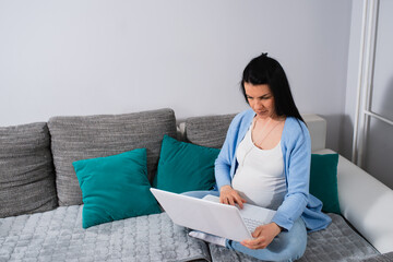 Freelence pregnant white woman sits in living room with crossed legs and holds laptop in her lap. She wears earphones. Black-haired female types on keyboard with her right hand and looks at the screen
