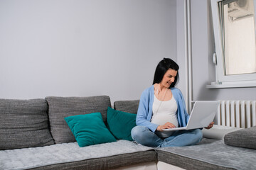 A child-bearing caucasian brunette sits on a sofa in the corner of the living room with crossed legs and holds a white laptop in her lap. She types on the keyboard and looks at the screen. 