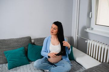 A beautiful, good-looking pregnant woman with a naked belly sits on sofa with crossed legs and combs her long, black hair. She looks aside and caresses her hair with left hand. She is in bright room. 