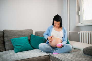 A child-bearing Caucasian female sits on a couch with crossed legs and smears her belly with a greasy cream. A brunette woman looks at her naked stomach and navel. A pink box of cream is in left hand.