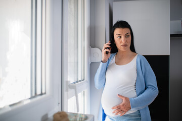 Unhappy pregnant caucasian brunette uses mobile phone and holds it on her ear while she seriously looks at her left side somewhere. She holds her big tummy with her left hand. Woman stands near door. 