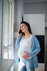 A good-looking caucasian pregnant woman is in the apartment. A beautiful female speaks on her mobile phone while she looks through the door and touches her big stomach. Her outfit is casual. 