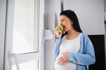 A healthy caucasian pregnant woman stands and looks through the window. She bites a green fresh apple and touches her belly with her left hand.  