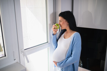 Young caucasian pregnant woman smells green apple while her eyes are closed. Black-haired woman is turned to bright door. She holds apple on her right hand and supports her tummy with her left hand.