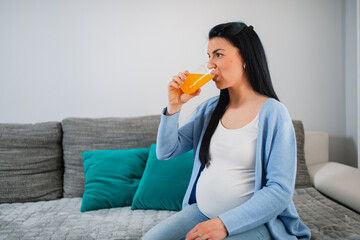 A healthy pregnant caucasian woman drinks orange mixed fresh juice and takes in necessary vitamins. Black-haired female sits on sofa in living room. She holds glass of orange juice in her right hand. 