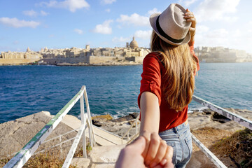 Follow me in Malta. Young carefree tourist woman in hat holds boyfriend hand and looking panoramic view of Valletta, Malta. Couple on summer holiday vacation. Traveling together. - obrazy, fototapety, plakaty